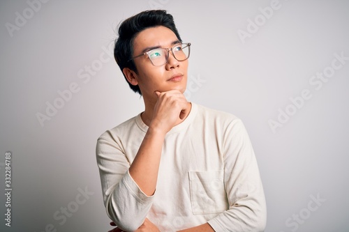 Fototapeta Naklejka Na Ścianę i Meble -  Young handsome chinese man wearing casual t-shirt and glasses over white background with hand on chin thinking about question, pensive expression. Smiling with thoughtful face. Doubt concept.