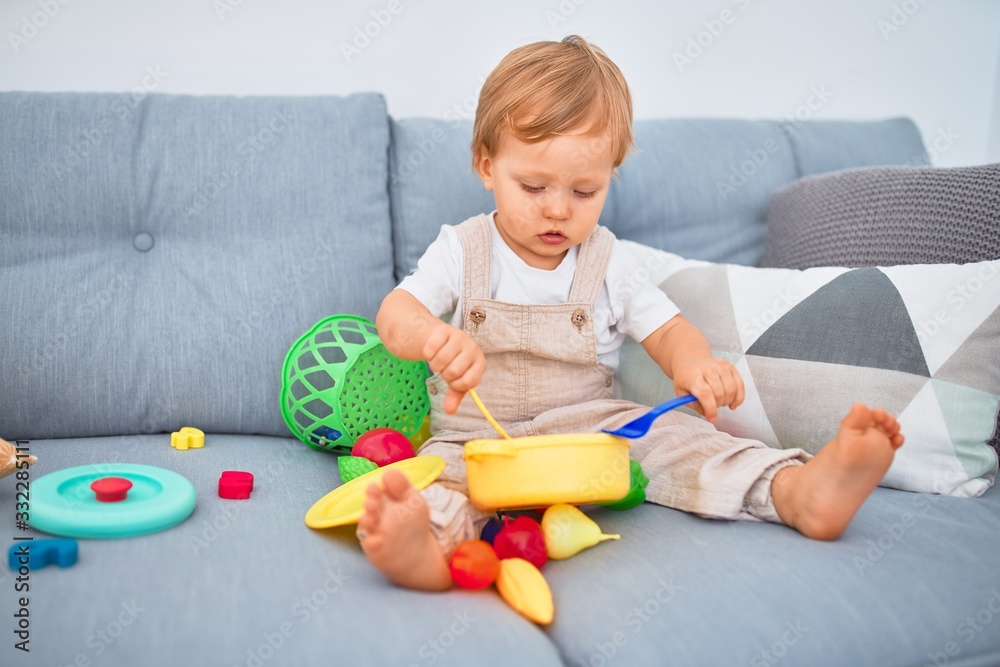 Adorable blonde toddler sitting on the sofa playing with plastic meals toys at home Adorable blonde toddler sitting on the sofa playing with plastic meals toys at home