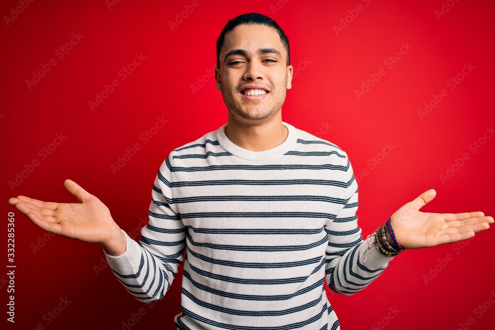 Young brazilian man wearing casual striped t-shirt standing over isolated red background smiling showing both hands open palms, presenting and advertising comparison and balance Young brazilian man wearing casual striped t-shirt standing over isolated red background smiling showing both hands open palms, presenting and advertising comparison and balance
