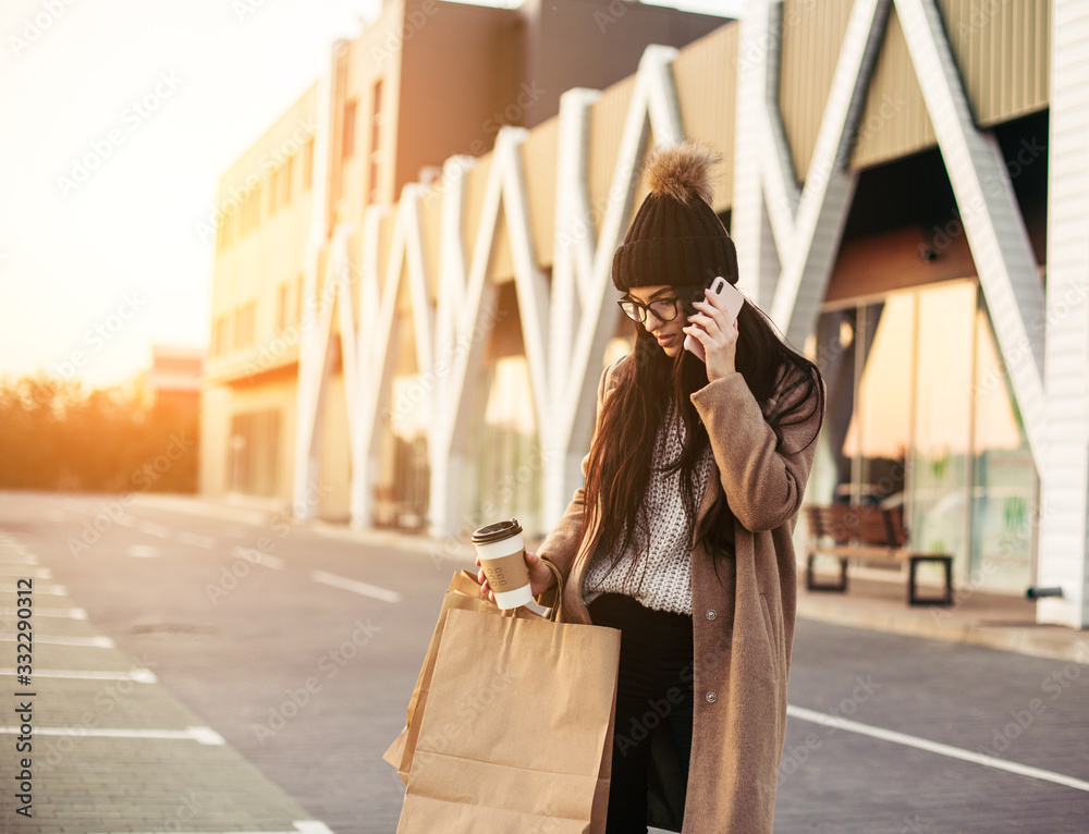 Young trendy woman blogger with shopping bag and smartphone near mall