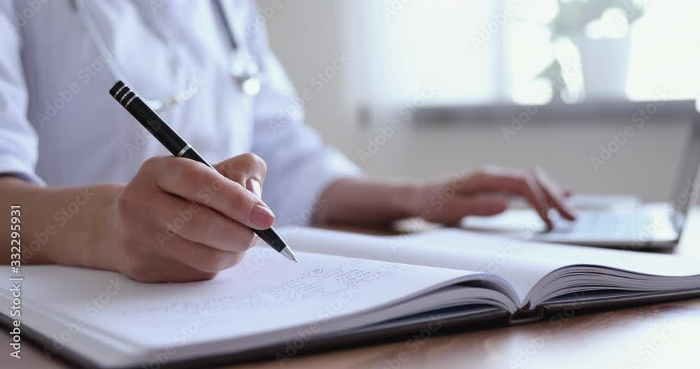 Close up view of female doctor holding pen making notes while doing online research. Professional physician wearing white uniform writing medical records or report in paper notebook sitting at desk. Close up view of female doctor holding pen making notes while doing online research. Professional physician wearing white uniform writing medical records or report in paper notebook sitting at desk.