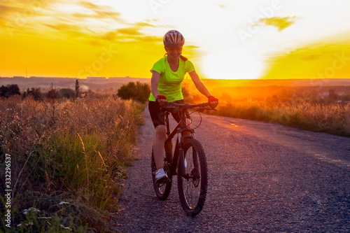 Beautiful girl cyclist riding a bike on the road towards the sunset. Nature and recreation. Hobbies and sports