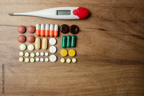 A lot of various pills  a body temperature digital thermometer on a wooden table in a household. Medical protective gear. A thermometer and medications against the COVID-19 virus other diseases.