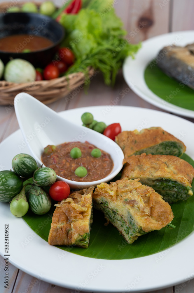 Shrimp paste, along with omelette, Cha-om and eggplant, placed in a white plate on a wooden floor. Shrimp paste, along with omelette, Cha-om and eggplant, placed in a white plate on a wooden floor.