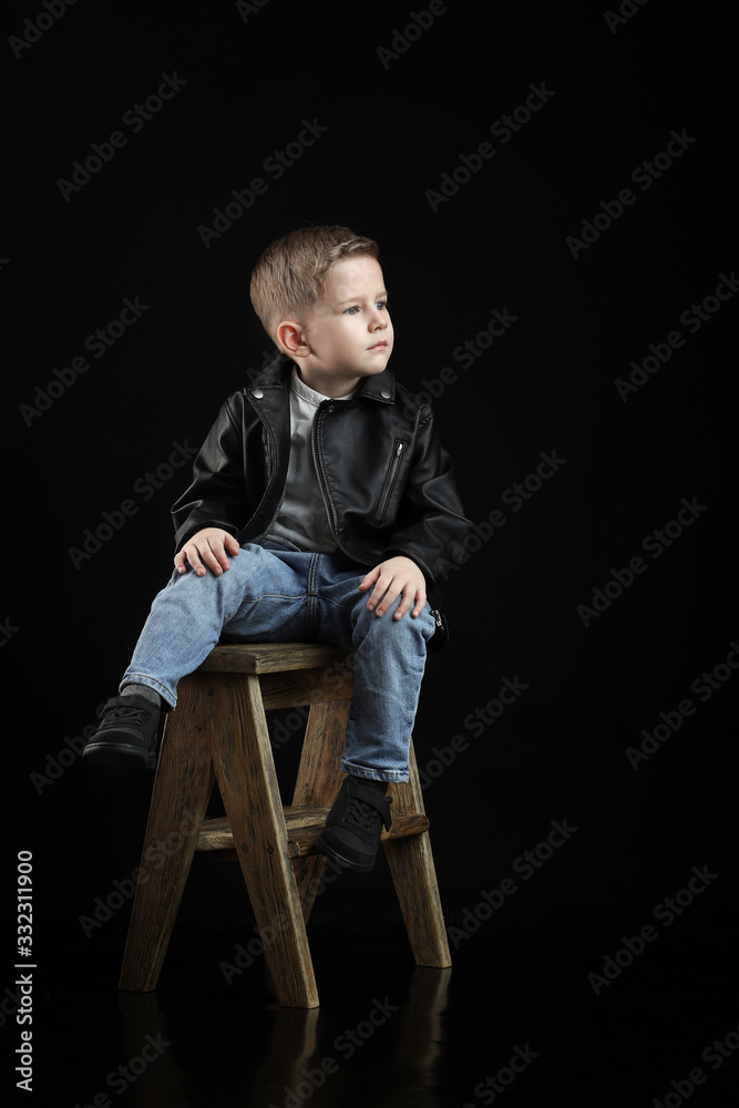 a fashionable boy in a leather black jacket and jeans sits on a chair on a black background with reflection on the floor. a fashionable boy in a leather black jacket and jeans sits on a chair on a black background with reflection on the floor.