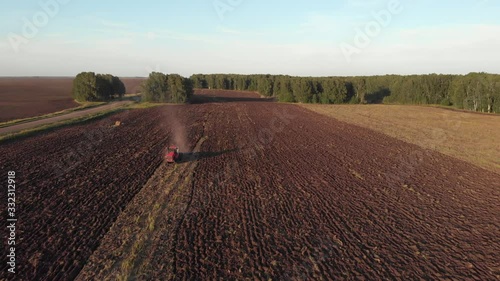 Drone camera moves towards a red tractor riding in a field and plowing fertile soil in the countryside