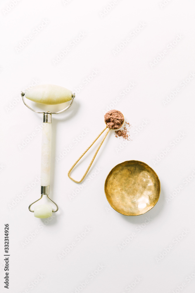 Quartz jade roller and scoop of herbal facial powder in spoon with brass dish on white surface / Holistic wellness and self care concept