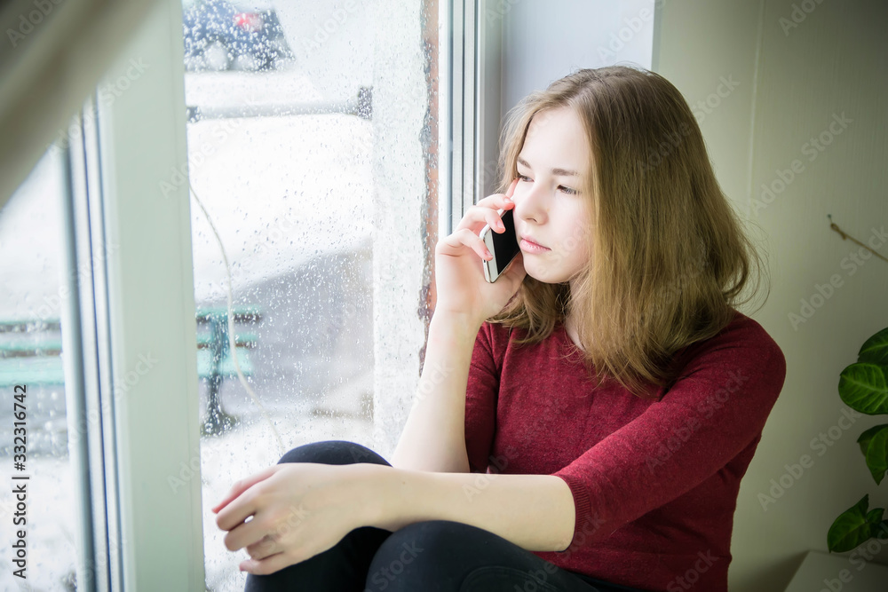 A teenage girl with long hair sits on a windowsill and talks, calls on her cell phone. It is raining outside the window.