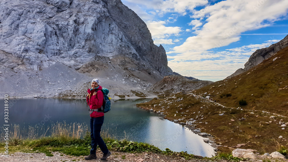 A woman in hiking outfit standing by the Wolayer Lake in Austrian Alps. There is massive, rocky mountain on the other side of the lake. New day beginning. Soft reflections in the lake. Happiness