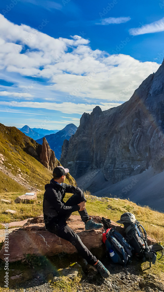 Fototapeta premium A man in hiking outfit resting and enjoying the view on Austrian Alps in autumn. There is massive, rocky mountain behind him. New day beginning. Contemplation and meditation. Calm state of mind.