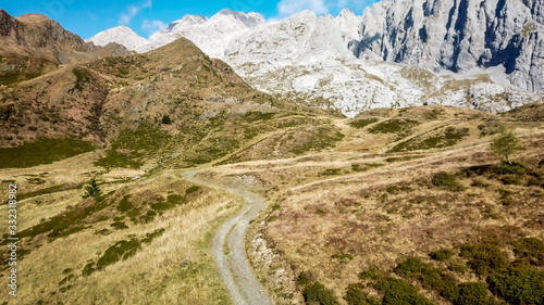 A drone shot of a pathway in Alps. The road is curvy, but steadily leading to the top. Golden autumn vibes. There is a tall, rocky mountain in the back. High Alpine mountain climbing