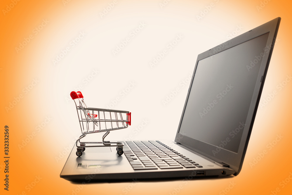 A laptop with a toy shopping cart sitting on the keyboard. Isolate on an orange background. Copy space. Side view. Concept of online shopping and modern technologies