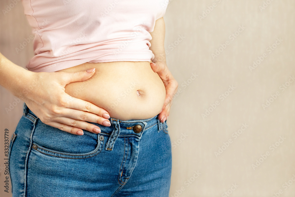Woman in jeans and a light shirt is standing sideways and holding hands squeezes belly fat on light background, copy space. The concept of overweight, weight loss, diet, obesity, junk food. Woman in jeans and a light shirt is standing sideways and holding hands squeezes belly fat on light background, copy space. The concept of overweight, weight loss, diet, obesity, junk food.