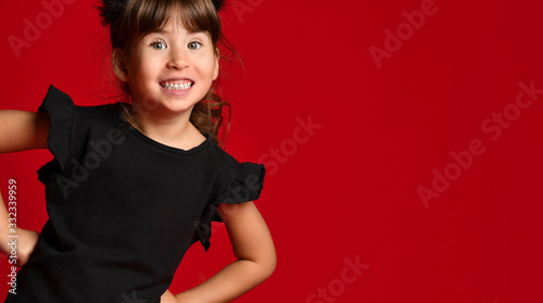 Cute brunette girl in a black T-shirt on a red background. The baby put her hands on her hips and laughs smiling at the camera.