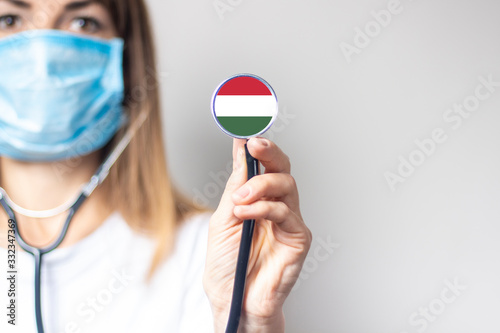 female doctor in a medical mask holds a stethoscope on a light background. Added flag of Hungary. Concept medicine, level of medicine, virus, epidemic