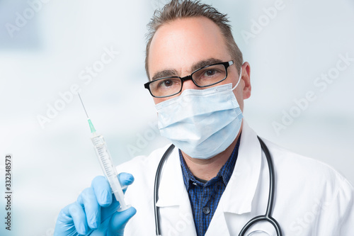 male doctor with medical face mask and medical gloves holding a syringe in front of a clinic room photo