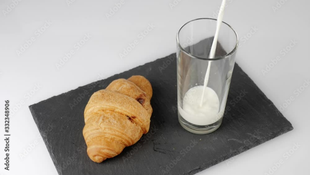 Fresh white milk is poured into a transparent glass near a croissant with chocolate on a black board on a white table. Concept of stay at home and eat healthy food. Delicious breakfast. Top view. 4K Fresh white milk is poured into a transparent glass near a croissant with chocolate on a black board on a white table. Concept of stay at home and eat healthy food. Delicious breakfast. Top view. 4K