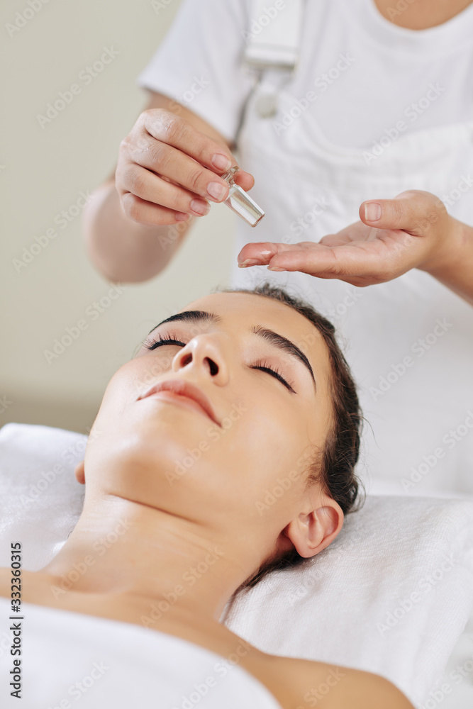 Beautician applying massage oil on face of young spa salon client