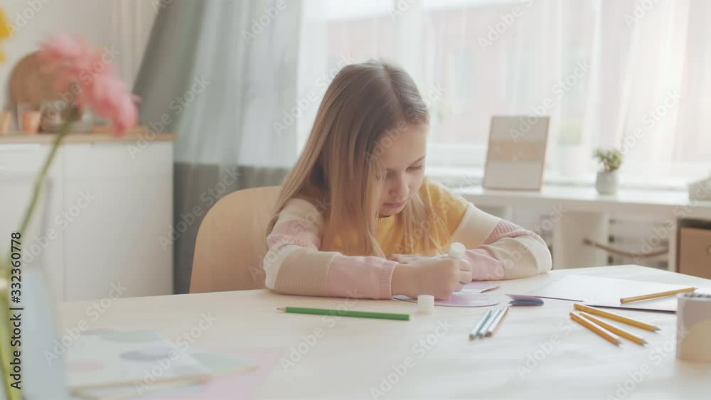 Lockdown of wonderful Caucasian girl with blond hair sitting at table and sticking paper details on handcrafted card using glue stick Lockdown of wonderful Caucasian girl with blond hair sitting at table and sticking paper details on handcrafted card using glue stick
