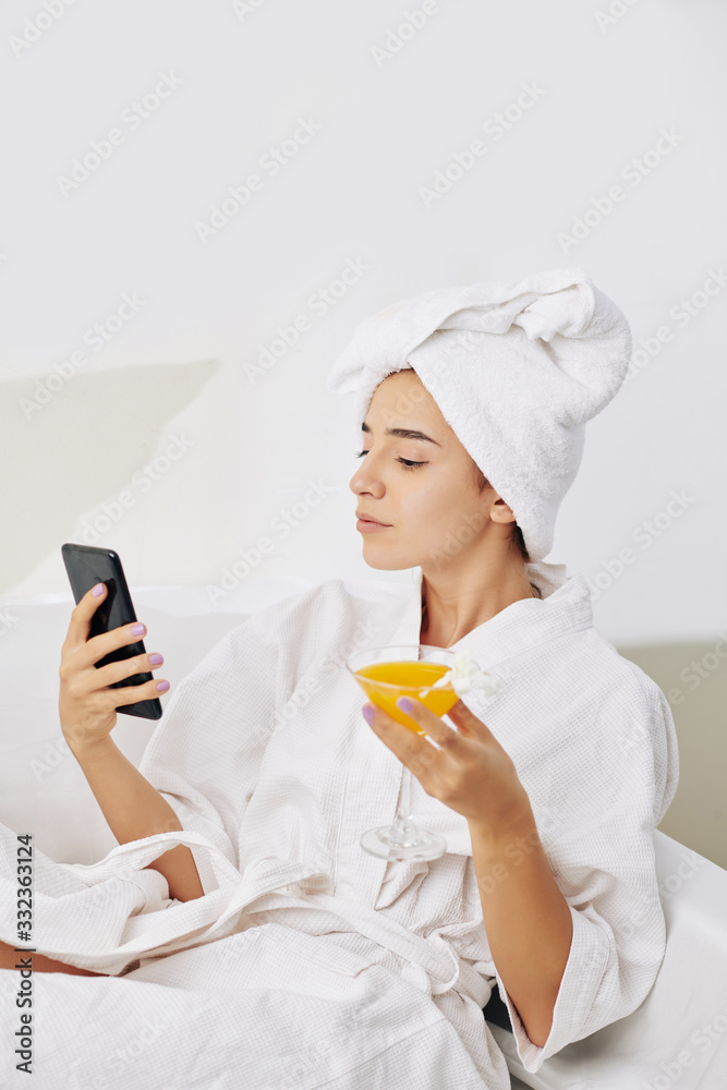 Beautiful young woman drinking sweet cocktail and checking social media on smartphone after taking bath