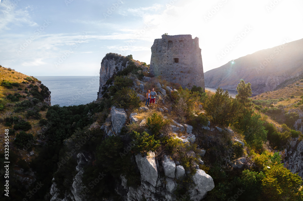 A couple of tourists on the background of the old tower, fortification. Mountains, rocky shores. Good sunny weather in summer. Recreation and tourism. Nerano, Naples, Italy