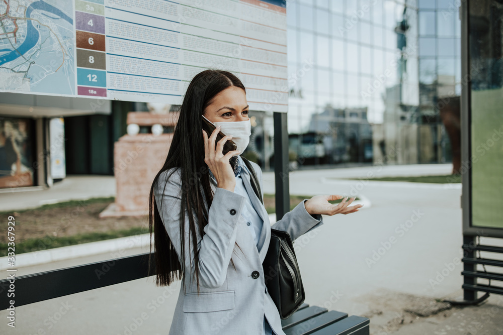 Elegant business woman with protective mask sitting alone on empty street and waiting for bus or taxi transport while using phone. Corona virus or Covid-19 lifestyle concept. Elegant business woman with protective mask sitting alone on empty street and waiting for bus or taxi transport while using phone. Corona virus or Covid-19 lifestyle concept.