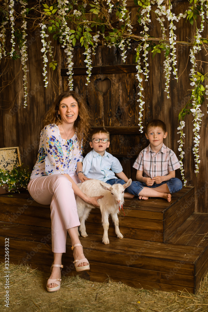 sitting young mother and two her sons kids with little white goat on dark wooden background with flowers and hay