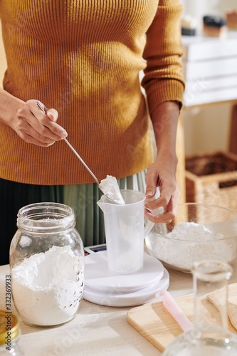 Cropped image of woman putting soda lye powder in plastic jar and measuring it on electronic scale when making soap mixture