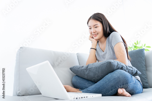 Asian young woman happy and smile using laptop connect internet for working at home and sitting on gray color sofa decoration by plants tree on white background