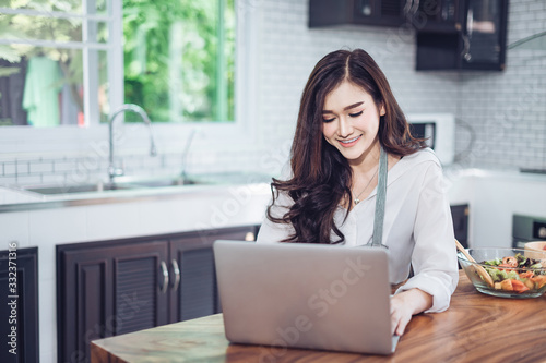 Image of young pretty woman standing in kitchen using computer laptop and cooking with the tomatoes and avocado.