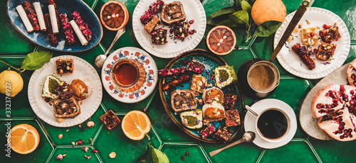 Flat-lay of variety of Turkish traditional lokum sweet delight with Turkish coffee in cups, tea in tulip glass and fresh fruits over green Moroccan tile table, top view. Middle East typical dessert