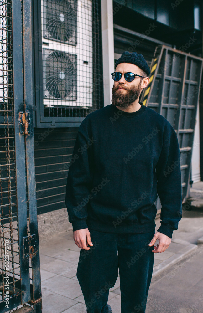 City portrait of handsome hipster guy with beard wearing black blank hoody or sweatshirt and hat with space for your logo or design. Mockup for print City portrait of handsome hipster guy with beard wearing black blank hoody or sweatshirt and hat with space for your logo or design. Mockup for print