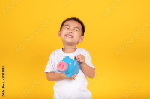 Happy Asian Little Children Boy saved a little money for future need wearing white T-shirt holding piggy bank and coin, Saving money since childhood on yellow background studio shot with copy space.