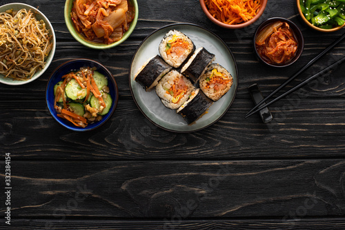 top view of plate with gimbap and chopsticks near tasty korean side dishes on wooden surface