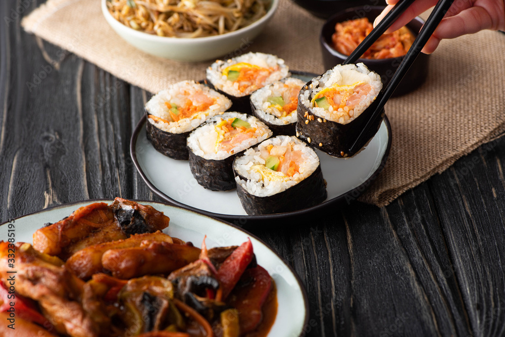 cropped view of woman holding chopsticks with rice roll near korean side dishes cropped view of woman holding chopsticks with rice roll near korean side dishes