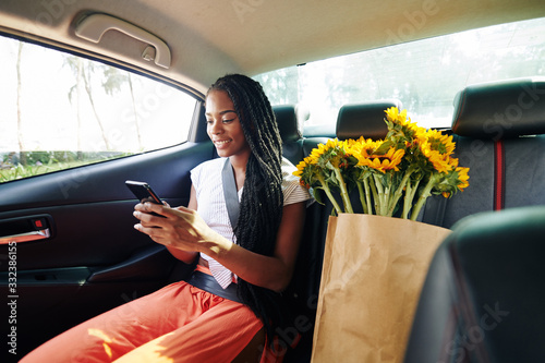 Beautiful positive young Black woman riding in taxi after supermarket and reading text messages from friends