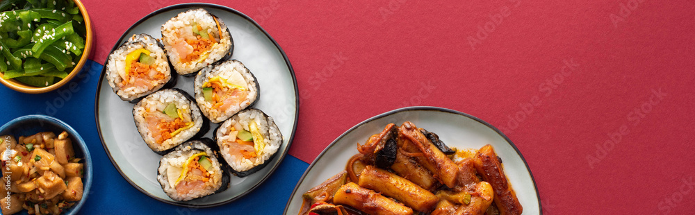 panoramic shot of plates with tasty gimbap near korean side dishes on blue and crimson panoramic shot of plates with tasty gimbap near korean side dishes on blue and crimson