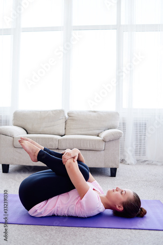 Portrait of young woman practicing apanasana pose on mat at home indoor, copy space. Apanasana. Knees to chest pose. Practicing yoga. Wellness and healthy lifestyle photo