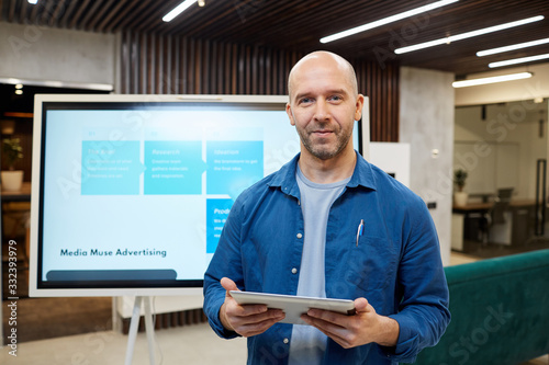 Waist up portrait of bald adult man smiling at camera and holding tablet while standing against digital board during marketing presentation, copy space