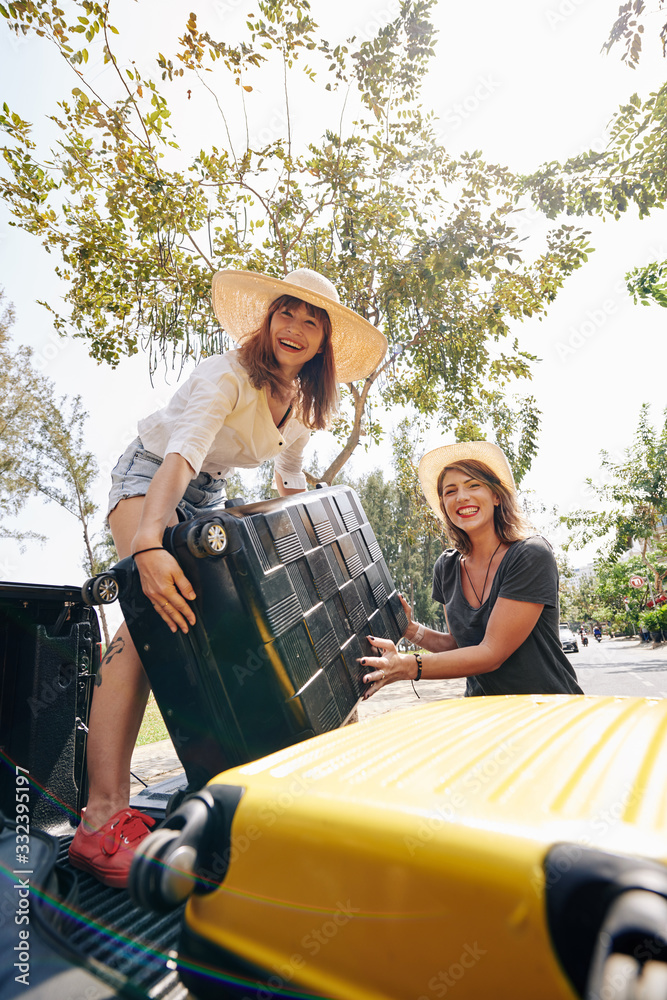 Happy pretty young female travelers putting heavy suitcases in car trunk