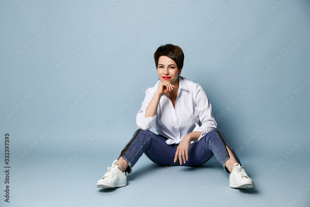 Smiling brunette woman in stylish blue jeans and white shirt is sitting on the floor with her legs apart looking at us