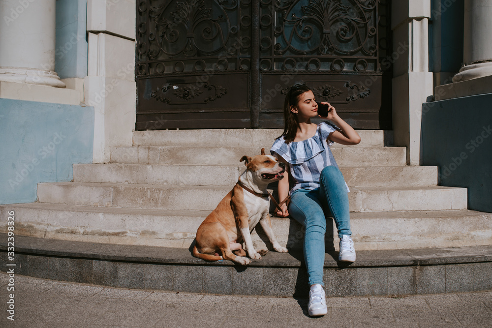  A young beautiful girl is talking on the phone next to her dog. Girl and dog on the stairs of the building