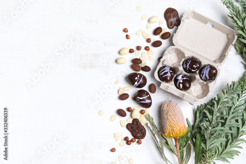 A selection of vegan chocolate Easter eggs in a recycled egg carton, surrounded by shaved Almonds, hazelnuts, Australian native Banksia flower on a rustic white background.