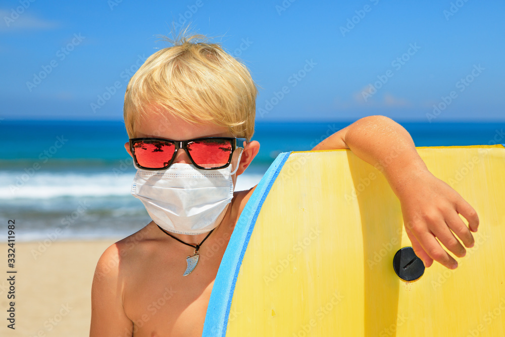Young surfer wearing sunglasses, protective mask on sea beach with body board. Summer tours, cruises cancellation due to coronavirus COVID-19 epidemic. Safe travel destinations for family vacation.