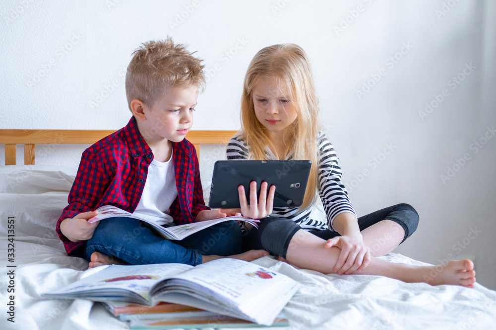Distance learning online education. Schoolboy and girl studying at home with digital tablet laptop notebook and doing school homework. Sitting on bed with training books