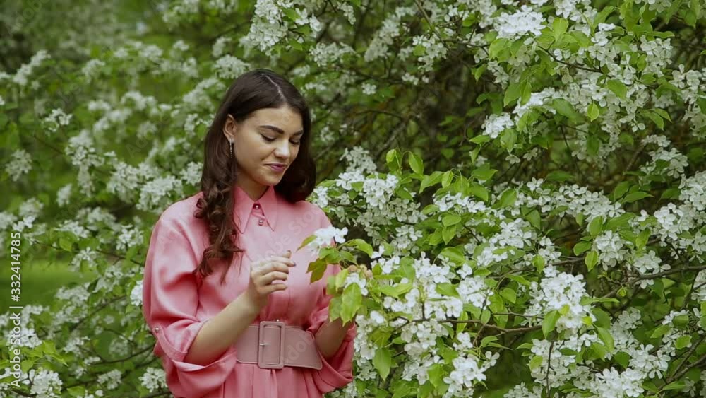 Beautiful stylish brunette girl with long hair in pink dress is sniffing the flowers in the blooming forest Beautiful stylish brunette girl with long hair in pink dress is sniffing the flowers in the blooming forest