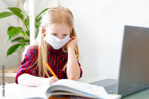 Distance learning online education. Sickness schoolgirl in medical mask studying at home with digital tablet in hand and doing school homework. Training books and notebooks on table photo