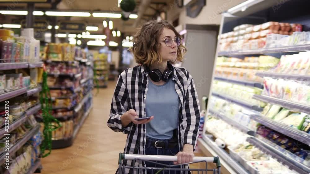 Shopping, technology, sale, consumerism and people concept - woman with smartphone and headphones on neck walking with trolley at supermarket or store, looking at mobiles screen. Front view