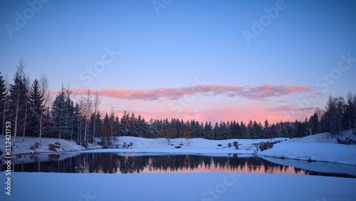 Winter landscape. The forest lake is partially covered with ice, surrounded by rocks, shrubs and spruces against the evening sky painted by the rays of the setting sun. © Александр Овсянников