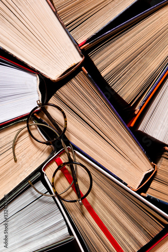 Many multicolored thick open books stand on a dark background. On the books are old round glasses